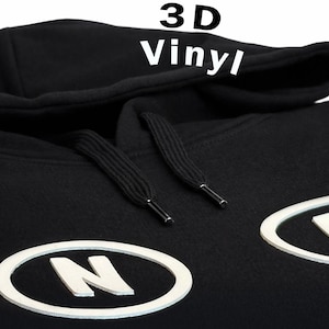 May include: Black hoodie with white '3D Vinyl' text on the hood. The hoodie features white oval patches with the letters 'N' and 'D'. The hood has black drawstrings with silver tips.
