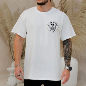 May include: White t-shirt with a circular graphic on the chest. The graphic features a skeleton with sunglasses giving a thumbs-up, surrounded by the words "COOL DADS CLUB". The shirt has short sleeves and a crew neck.