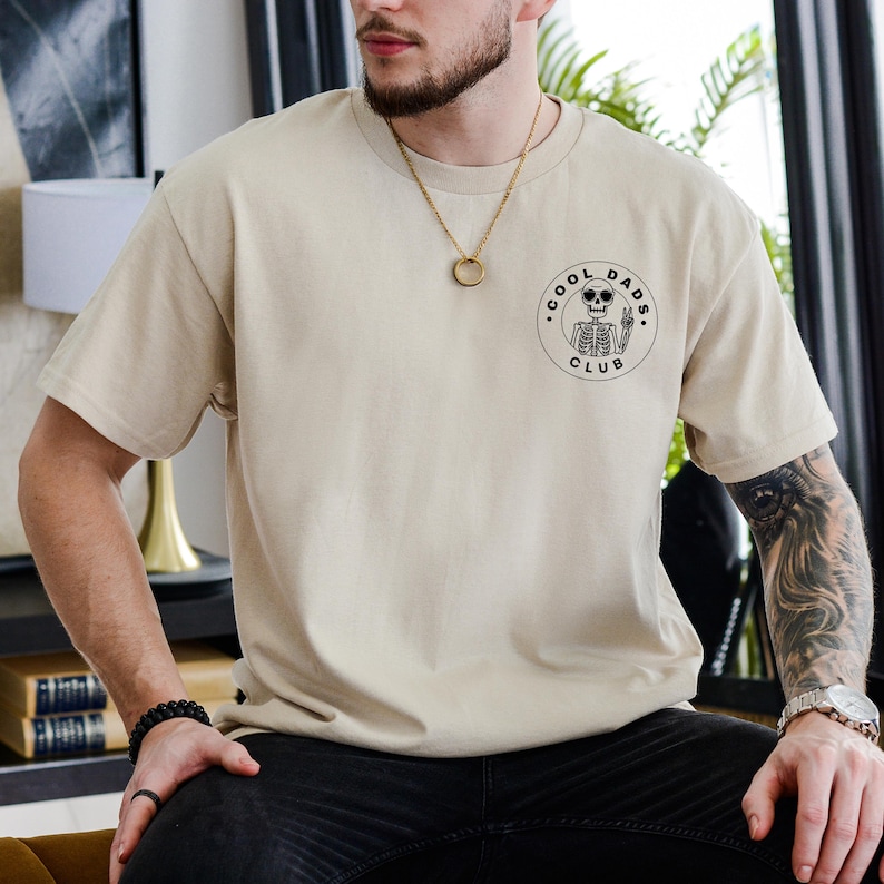 May include: Beige short-sleeved t-shirt with a round black graphic on the left chest. The graphic features a skeleton wearing sunglasses and the words "COOL DADS CLUB" in a circular design. The shirt is a neutral color.