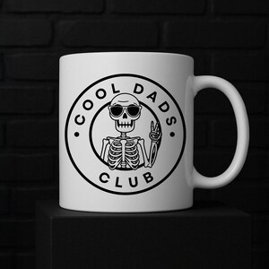 May include: White ceramic mug with a black graphic of a skeleton wearing sunglasses and giving a peace sign. The text "COOL DADS CLUB" is printed around the skeleton in a circular design. The mug has a curved handle.