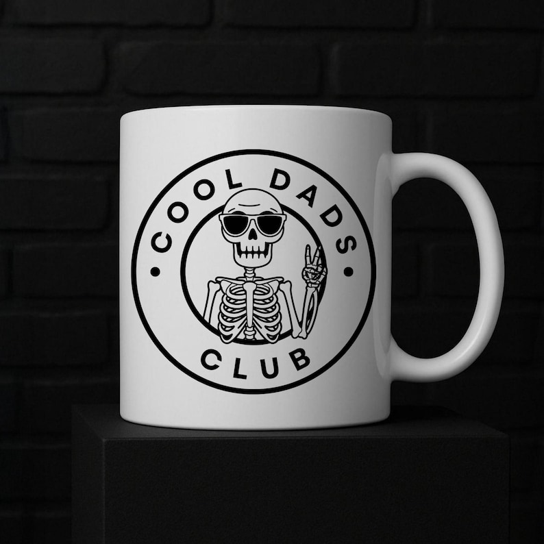May include: White ceramic mug with a black graphic of a skeleton wearing sunglasses and giving a peace sign. The text "COOL DADS CLUB" is printed around the skeleton in a circular design. The mug has a curved handle.