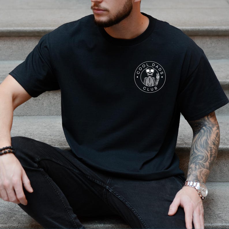 May include: Black t-shirt with a white circular graphic that reads "COOL DADS CLUB" around a skeleton holding a beer. The shirt is a crew neck and has short sleeves.