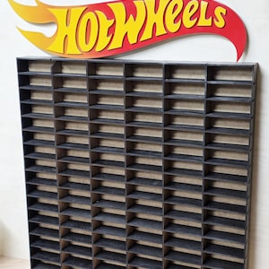 May include: A black wooden display shelf with multiple compartments for toy cars. The Hot Wheels logo, in yellow and red, is above the shelf. The shelf is designed to hold and organize small toy cars.