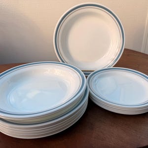 May include: A collection of white ceramic dishes with a blue and gray border. The set includes several bowls and plates, stacked and arranged on a wooden table. The dishes have a simple, classic design, suitable for everyday use.