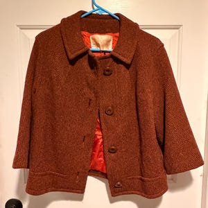 Könnte beinhalten: Eine Vintage-Jacke aus rostfarbener Wolle mit klassischem Kragen und drei Knöpfen. Die Jacke hat Dreiviertelärmel und ein leuchtend orangefarbenes Futter. Die Jacke hängt an einem blauen Kleiderbügel vor einer weißen Tür.