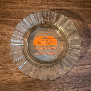 May include: Clear glass ashtray with a fluted rim. The center features the orange logo of Howard Johnson's restaurants and motor lodges. The ashtray is circular and sits on a wooden surface.