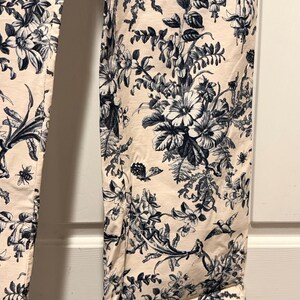 May include: Cream-colored pants with a detailed blue floral pattern. The design includes various flowers, leaves, and insects. The pants have a classic toile de jouy style, with a rolled-up hem.