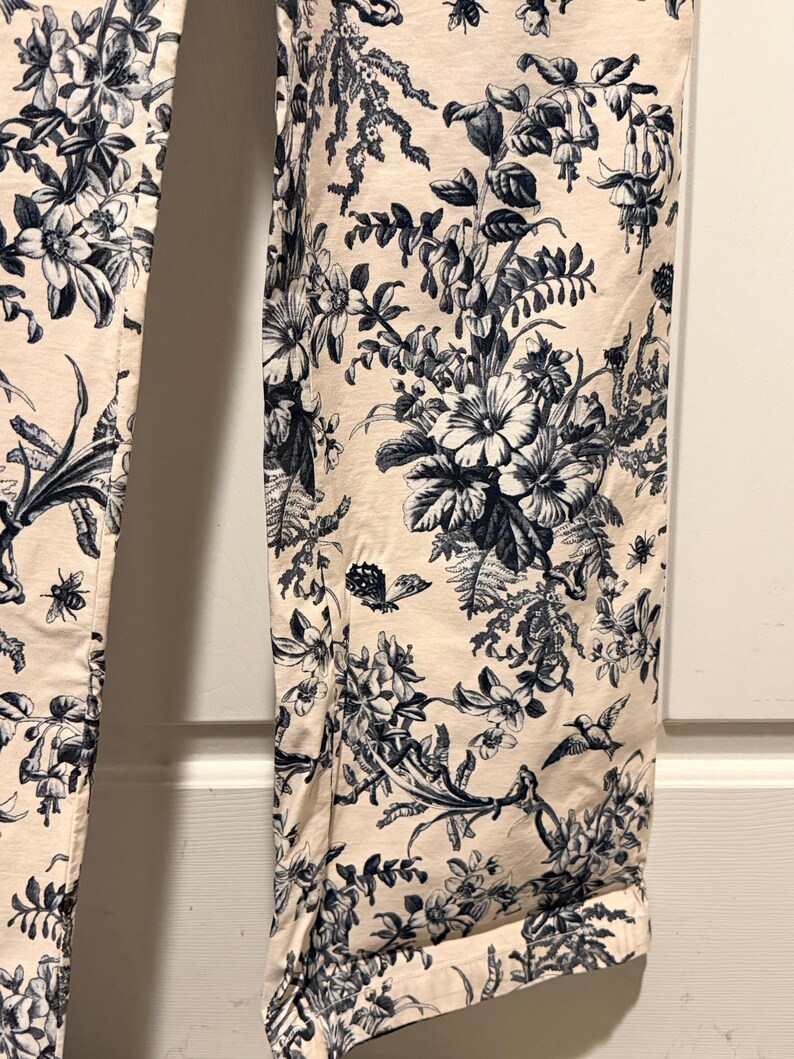May include: Cream-colored pants with a detailed blue floral pattern. The design includes various flowers, leaves, and insects. The pants have a classic toile de jouy style, with a rolled-up hem.