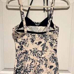 May include: A cream-colored jumpsuit with a black halter top and straps. The jumpsuit features a floral pattern in navy blue. The garment is hanging on a white hanger against a white door.