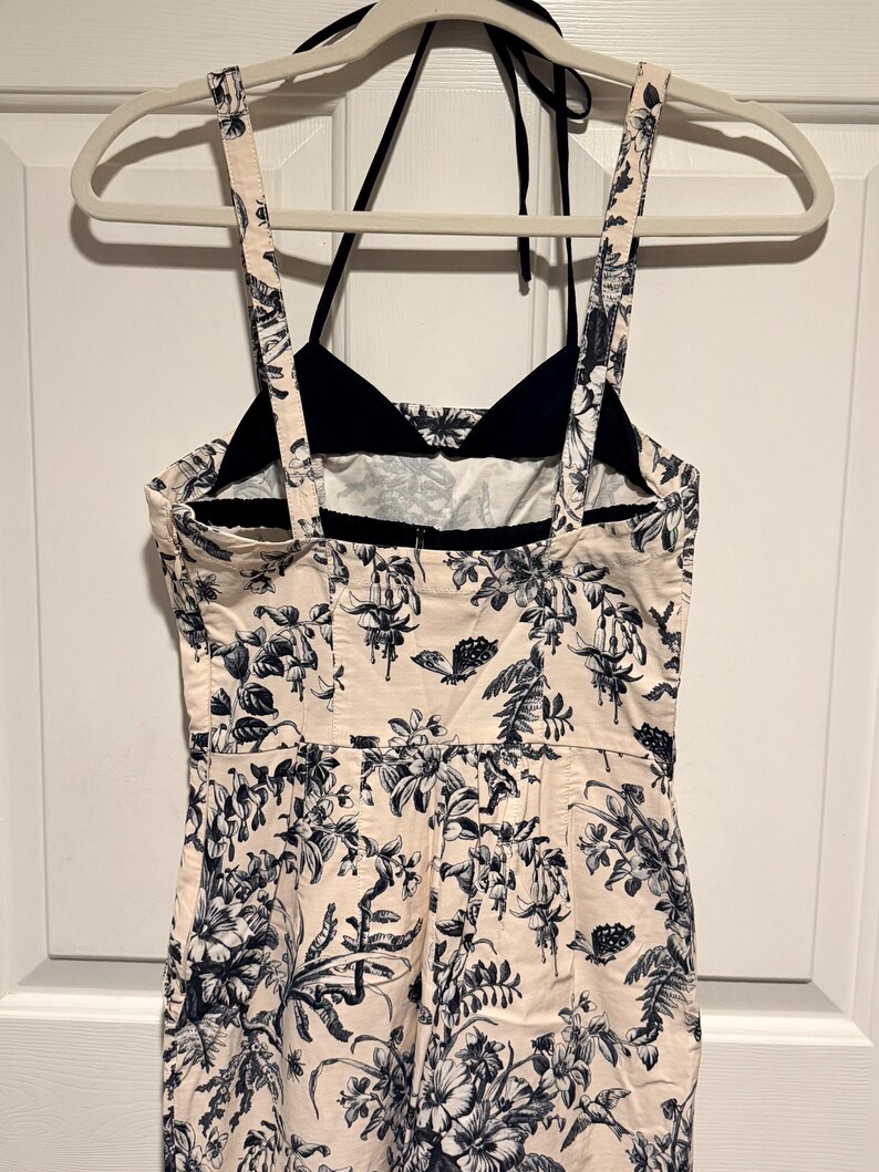 May include: A cream-colored jumpsuit with a black halter top and straps. The jumpsuit features a floral pattern in navy blue. The garment is hanging on a white hanger against a white door.