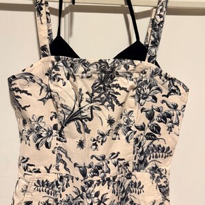 May include: A cream-colored jumpsuit with a black and white floral pattern. The jumpsuit has a halter top and wide straps. The floral design includes flowers, leaves, and insects. The jumpsuit is hanging on a white hanger.
