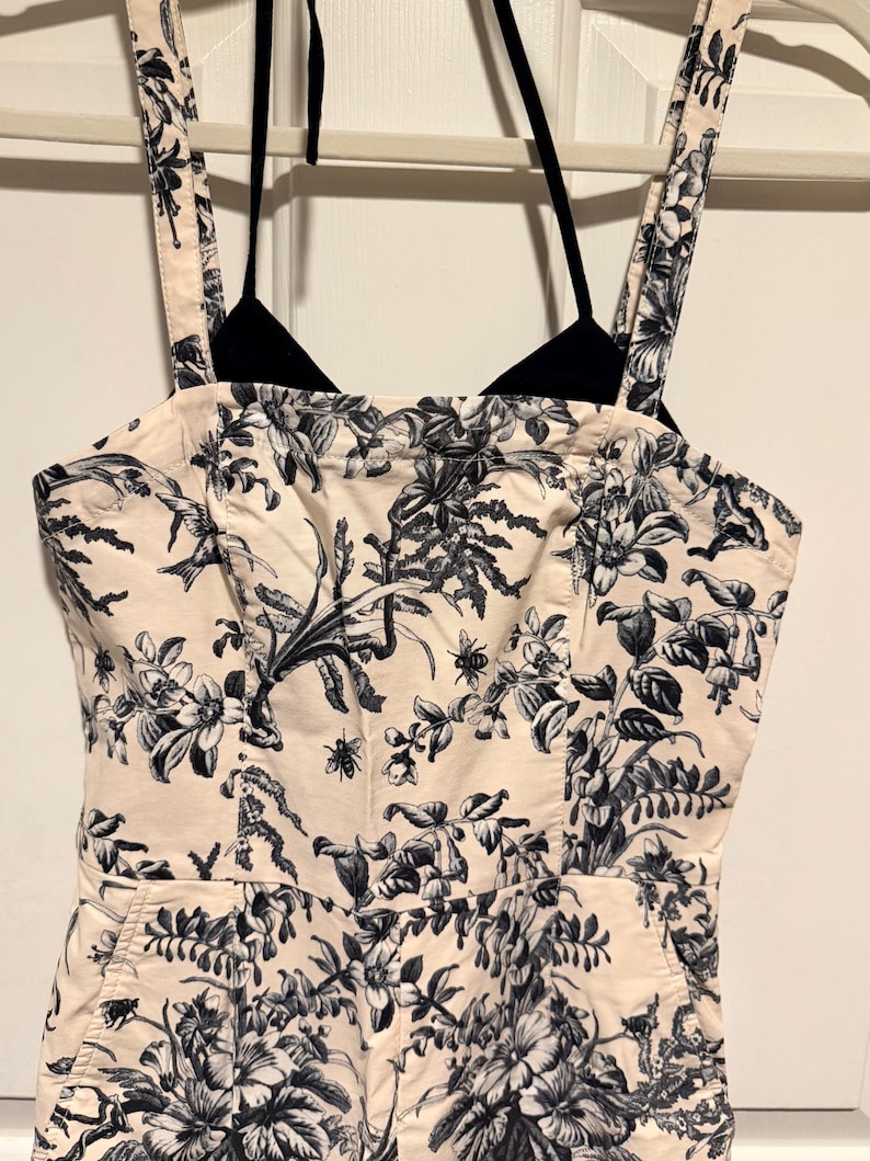 May include: A cream-colored jumpsuit with a black and white floral pattern. The jumpsuit has a halter top and wide straps. The floral design includes flowers, leaves, and insects. The jumpsuit is hanging on a white hanger.
