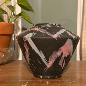 Puede incluir: Un jarrón de cerámica negro con forma geométrica. Está decorado con salpicaduras de pintura rosa y blanca. El jarrón está sobre una superficie de madera, con una planta en maceta al fondo.