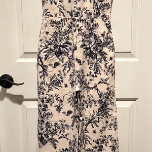 May include: A cream-colored jumpsuit with a black floral pattern. The jumpsuit has a halter neckline, a fitted bodice, and wide legs. It is hanging on a white hanger against a white door.