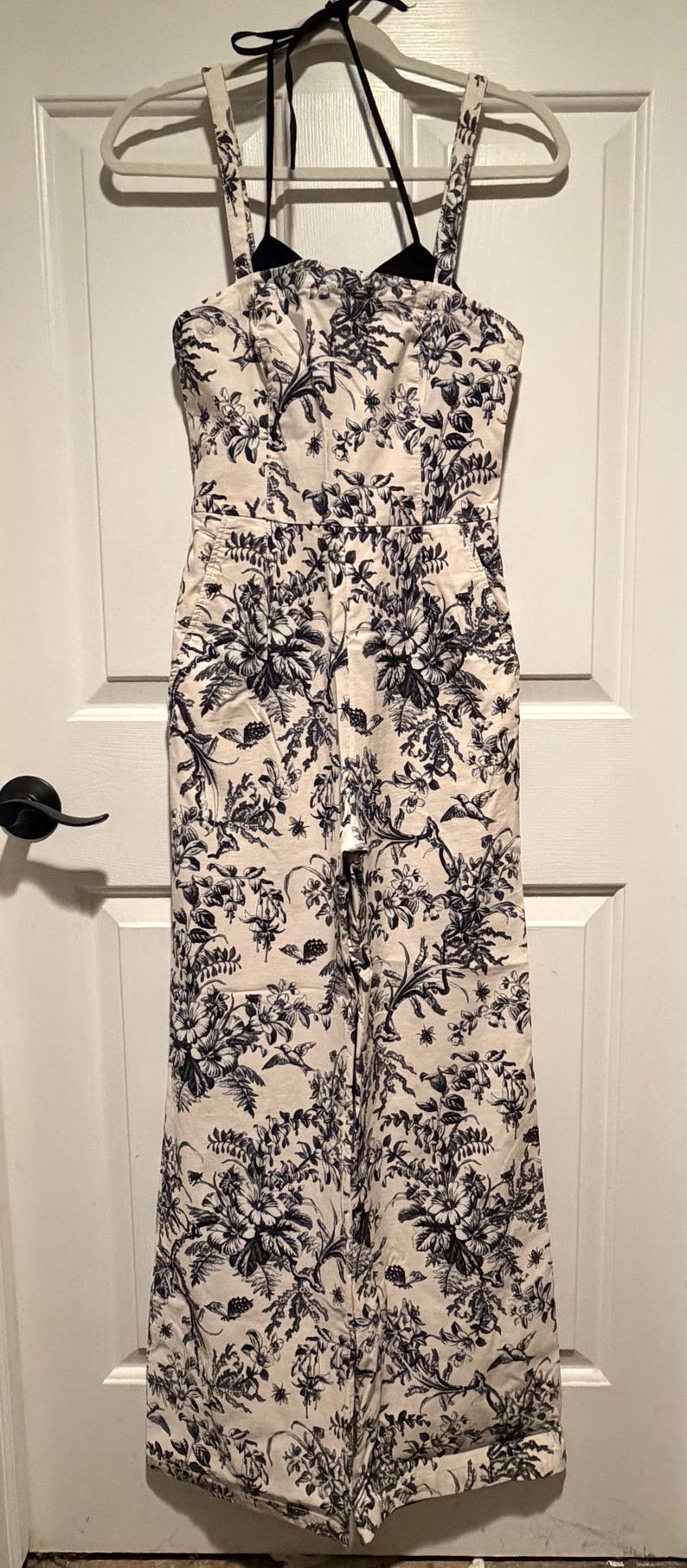 May include: A cream-colored jumpsuit with a black floral pattern. The jumpsuit has a halter neckline, a fitted bodice, and wide legs. It is hanging on a white hanger against a white door.