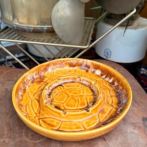 May include: A vintage, round ashtray with a textured, honeycomb-like pattern in shades of brown and yellow. The ashtray has a raised center and a wide, flat rim. The design is reminiscent of mid-century modern decor.