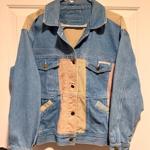 May include: A vintage denim jacket with a patchwork design. The jacket features a light blue denim body with tan and pink panels down the front. It has two chest pockets, button closures, and a classic collar.