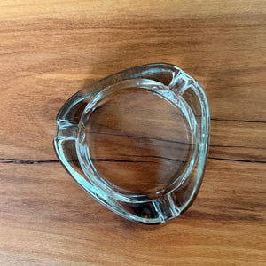 May include: Clear glass ashtray with a triangular shape and a circular indentation in the center. The ashtray has a smooth, transparent appearance and is designed with grooves for resting cigarettes. The ashtray is on a wooden surface.