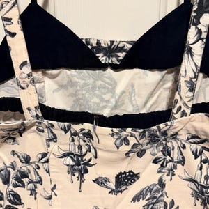 May include: A close-up of a cream-colored top with a black floral and butterfly print. The top has black straps and a black bodice. The floral print includes detailed flowers and leaves.