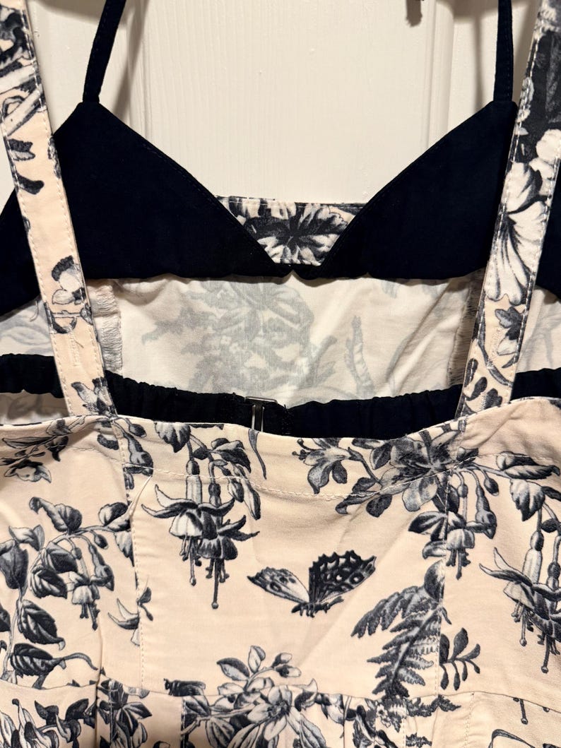 May include: A close-up of a cream-colored top with a black floral and butterfly print. The top has black straps and a black bodice. The floral print includes detailed flowers and leaves.