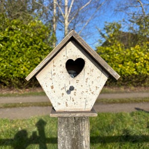 Puede incluir: Una casita de pájaros blanca con una abertura en forma de corazón. La casita de pájaros está montada en un poste de madera y tiene un aspecto desgastado y envejecido.