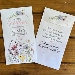 May include: A white card with a floral design and the text "a little Wildflower is on her way Kelsey's Baby Shower 08.19.2024". The card is next to a white envelope with the text "Plant these seeds and watch them bloom, just like the baby who will be here soon Thank you for sharing this special day with us!"