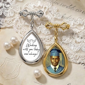 May include: Two teardrop-shaped memorial pendants. One is silver with the text "Walking with you today and always!" and angel wings. The other is gold, holding a photo of a person in a graduation cap and gown. Both have a bow-shaped pin.