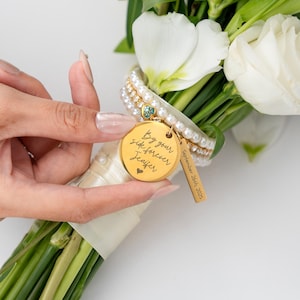 May include: A gold-colored round charm with the inscription "By your side forever Jennifer" and a heart symbol, attached to a bouquet of white flowers. A smaller rectangular charm reads "September 26th, 2025". The bouquet is adorned with pearls and a blue gemstone.