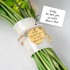 May include: A bouquet of flowers wrapped with a white ribbon, adorned with a gold charm bracelet. The bracelet features a round charm engraved with a message and a smaller rectangular charm. The text on the round charm reads "Emily, We love you so much Mom + Dad".