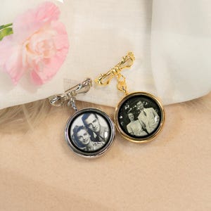 May include: Two silver and gold photo locket brooches with a black and white photo of a couple inside. The brooches have a bow-shaped pin and a chain attached to the locket.