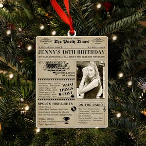 May include: A personalized Christmas ornament with a newspaper-style design. The ornament commemorates "Jenny's 18th Birthday" with a photo, date, and details about the year she was born. It includes sports highlights and radio hits.