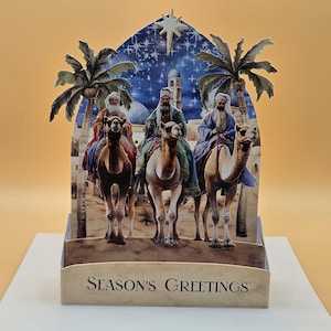 May include: A pop-up Christmas card featuring three figures riding camels, set against a backdrop of a starry night sky, palm trees, and Middle Eastern architecture. The card reads "SEASON'S GREETINGS".