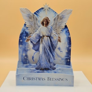 May include: A 3D Christmas card featuring an angel with large white wings and flowing blue robes. The angel stands in front of a blue backdrop with stars and a star at the top. The words "CHRISTMAS BLESSINGS" are printed at the bottom.