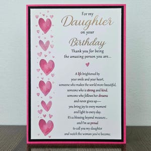 May include: A rectangular birthday greeting card with a pink border and black frame. The left side features a vertical column of watercolor-style pink hearts. The text reads: For my Daughter on your Birthday, Thank you for being the amazing person you are... A life brightened by your smile and your heart, someone who makes the world more beautiful, someone who is strong and kind. someone who follows her dreams and never gives up—you bring joy to every moment and light to every day. It's a blessing beyond measure... and I'm so proud to call you my daughter and watch the woman you've become.