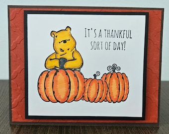 May include: A rectangular greeting card featuring a yellow bear sitting on a large orange pumpkin next to two smaller orange pumpkins. The text IT'S A THANKFUL SORT OF DAY! is printed in black. The card has a dark brown border and a textured red frame.
