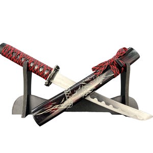 Op de afbeelding: Een miniatuur katana set met een zwarte schede en een rood-zwarte handgreep. Het lemmet is zilverkleurig en de schede is versierd met een wit ontwerp. De set wordt gepresenteerd op een zwarte standaard.