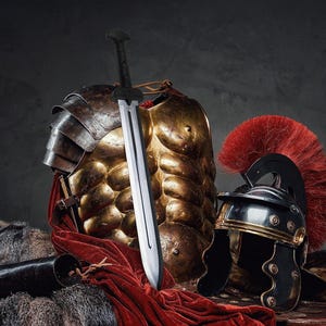 Könnte beinhalten: Eine Sammlung römischer Rüstungen und Waffen. Das Bild zeigt einen goldenen Brustpanzer, einen schwarz-goldenen Helm mit roter Feder und ein silbernes Schwert. Ein rotes Tuch und eine schwarze Lederrolle sind ebenfalls vorhanden.