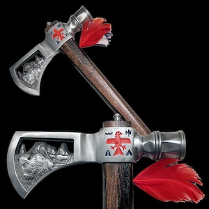 Könnte beinhalten: Zwei dekorative Tomahawks mit silberfarbenen Köpfen und dunklen Holzgriffen. Die Köpfe weisen detaillierte Schnitzereien eines Bisons und ein rotes Vogelsymbol auf. Rote Federn sind an den Griffen befestigt.