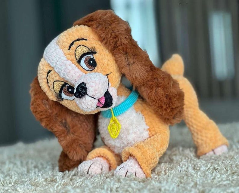 May include: A plush, handmade dog toy in shades of tan, brown, and white. The dog has large, expressive eyes, floppy ears, and a blue collar with a yellow tag. The toy is made of soft, textured material.