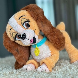 Puede incluir: Un juguete de peluche hecho a mano con forma de perro, en tonos beige, marrón y blanco. El perro tiene ojos grandes y expresivos, orejas caídas y un collar azul con una etiqueta amarilla. El juguete es de material suave y texturizado.
