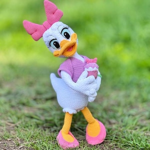 Puede incluir: Mu&ntilde;eca de ganchillo de Daisy Duck hecha a mano. La mu&ntilde;eca tiene un cuerpo blanco, un lazo rosa y una camisa morada. Sostiene una peque&ntilde;a magdalena. Los pies de la mu&ntilde;eca son amarillos con suelas rosas. El fondo es hierba verde.