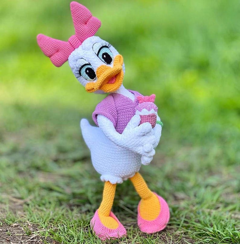 Puede incluir: Mu&ntilde;eca de ganchillo de Daisy Duck hecha a mano. La mu&ntilde;eca tiene un cuerpo blanco, un lazo rosa y una camisa morada. Sostiene una peque&ntilde;a magdalena. Los pies de la mu&ntilde;eca son amarillos con suelas rosas. El fondo es hierba verde.