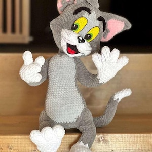 Puede incluir: Un peluche hecho a mano de Tom Cat de la caricatura Tom y Jerry. El gato es gris con patas blancas y un pecho blanco. Tiene orejas internas rosas, ojos amarillos y una boca roja. El gato está sentado sobre una superficie de madera.