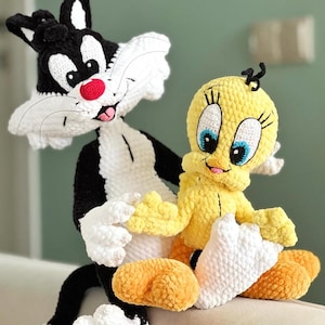 May include: Crocheted figures of Sylvester the Cat and Tweety Bird. Sylvester is black and white with a red nose and pink tongue. Tweety is yellow with blue eyes and white accents. Both figures are sitting on a light-colored surface.