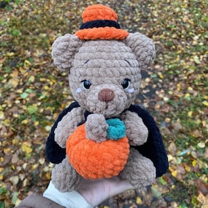 May include: A plush brown teddy bear wearing a black cape and an orange and black hat. The bear is holding an orange pumpkin with a green stem. The bear is a Halloween-themed plush toy.
