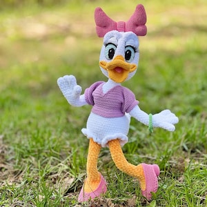 Puede incluir: Mu&ntilde;eca de ganchillo de Daisy Duck hecha a mano. La mu&ntilde;eca tiene cabeza, cuerpo y manos blancos, con un lazo, camisa y zapatos rosas. Las piernas de la mu&ntilde;eca son amarillas y tiene una pulsera verde. La mu&ntilde;eca est&aacute; sobre hierba verde.