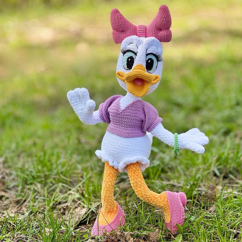 Puede incluir: Mu&ntilde;eca de ganchillo de Daisy Duck hecha a mano. La mu&ntilde;eca tiene cabeza, cuerpo y manos blancos, con un lazo, camisa y zapatos rosas. Las piernas de la mu&ntilde;eca son amarillas y tiene una pulsera verde. La mu&ntilde;eca est&aacute; sobre hierba verde.