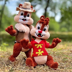 Puede incluir: Dos peluches de Chip y Chop hechos a mano con ganchillo. Una ardilla listada lleva un sombrero y una chaqueta marrones. La otra ardilla listada lleva una camisa roja con símbolos amarillos. Ambos tienen ojos grandes y expresivos y posan sobre hierba seca.