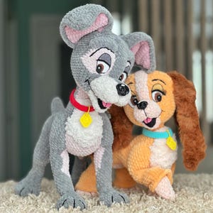 May include: Two plush crochet dog figures, one gray and one tan, inspired by a classic animated film. The gray dog has a red collar with a yellow tag, while the tan dog has a blue collar with a yellow tag. Both have smiling faces.