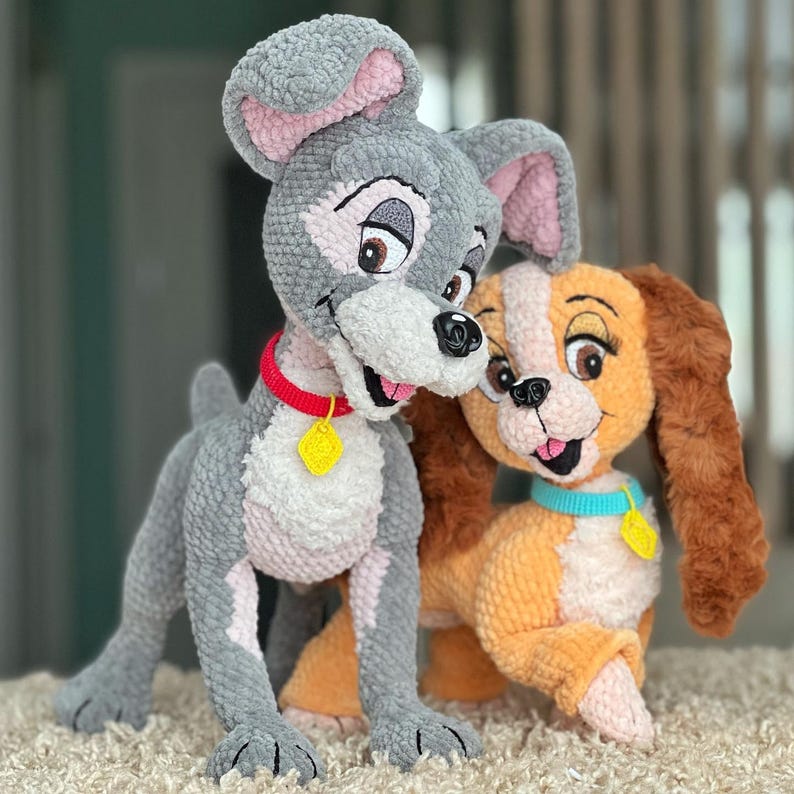 May include: Two plush crochet dog figures, one gray and one tan, inspired by a classic animated film. The gray dog has a red collar with a yellow tag, while the tan dog has a blue collar with a yellow tag. Both have smiling faces.